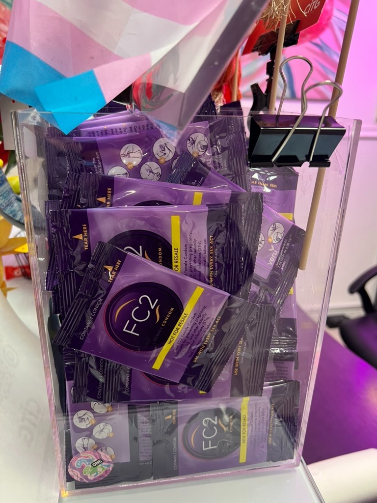 A clear acrylic holder on a desk filled with purple FC2 internal condom packets, with a transgender pride flag hanging behind it.