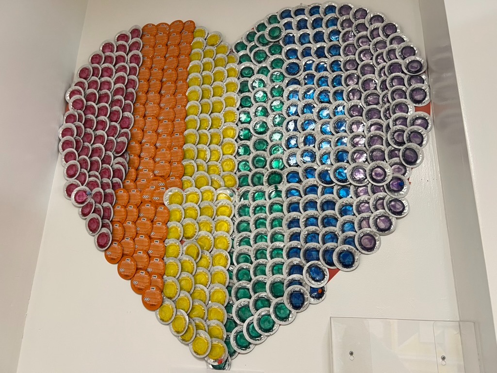 A large heart mounted on a white office wall, made of hundreds of foil-wrapped condoms arranged in rainbow stripes: pink, orange, yellow, green, blue and purple.