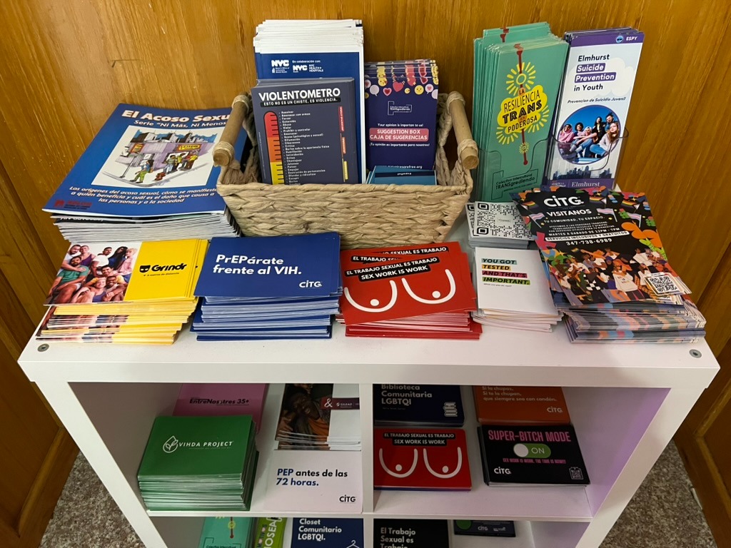 A white shelf stacked with free flyers and pamphlets in English and Spanish on HIV prevention, PrEP, PEP, sex workers' rights and trans resilience.